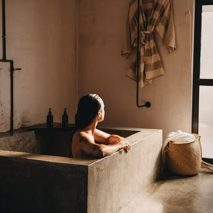 Person relaxing in a minimalist concrete bathtub, looking toward a window, with a robe hanging on the wall and a woven basket nearby.