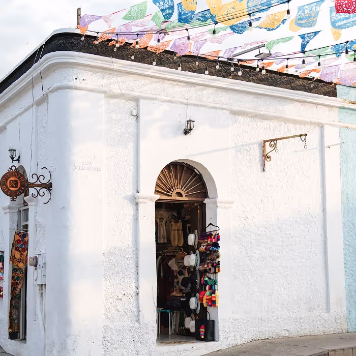 White stucco corner shop with an arched doorway displaying hats and colorful souvenirs, under strings of multicolored papel picado decorations.