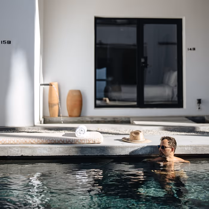 A man relaxes in a pool outside a modern white hotel room, wearing sunglasses as sunlight reflects on the water. A rolled towel and straw hat rest on the poolside ledge above him.