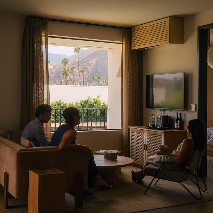 Three people relax in a cozy living room, chatting with drinks, as sunlight streams through a large window framing palm trees and mountains outside.