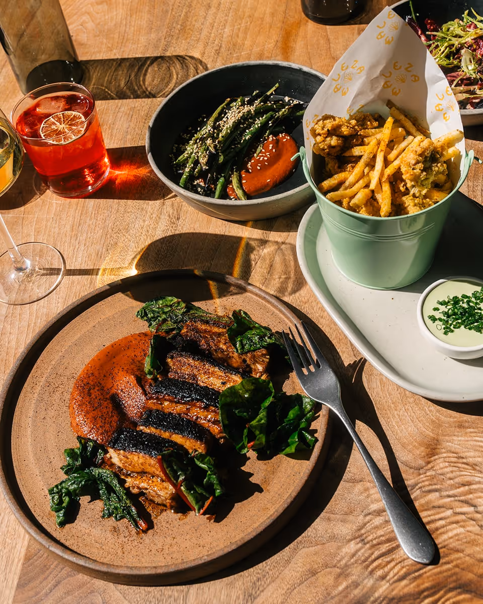 Assorted dishes on a wooden table including grilled meat with sauce, fries with dipping sauce, green beans, and colorful cocktails.