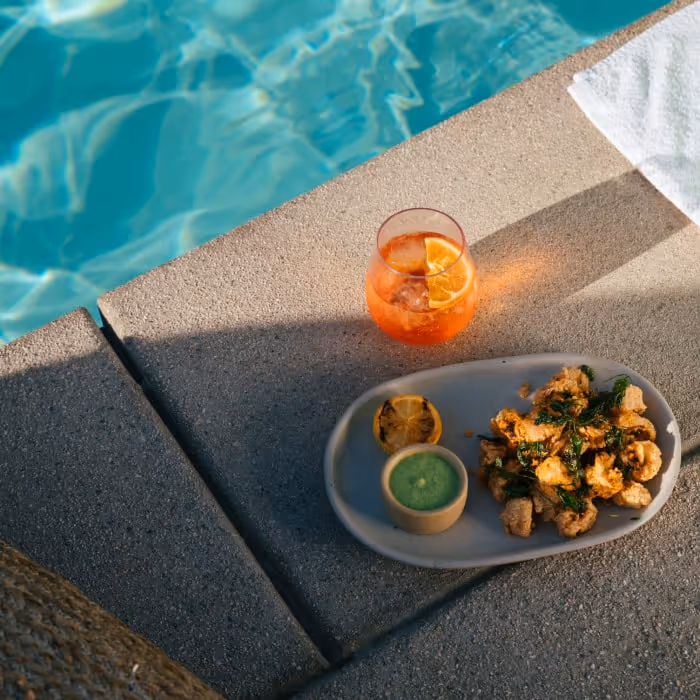 Colorful cocktail and plate of fried appetizers with dipping sauce by the poolside.