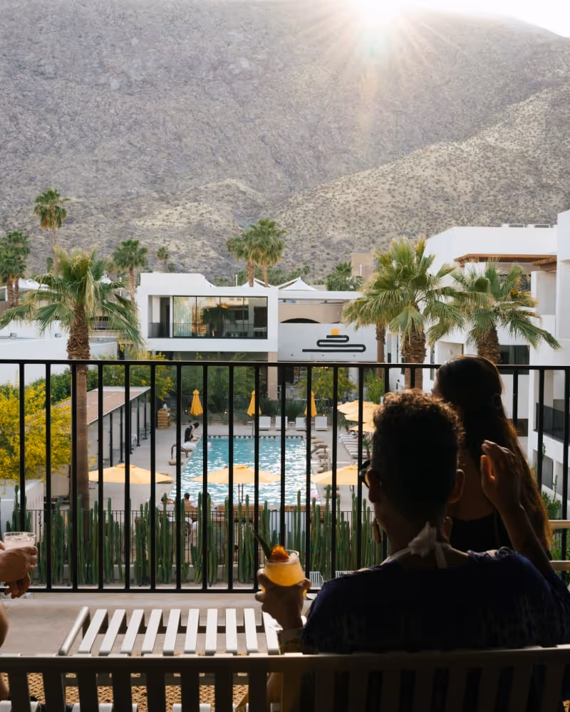 Guests relaxing on a balcony overlooking a pool with palm trees and mountain views at sunset.