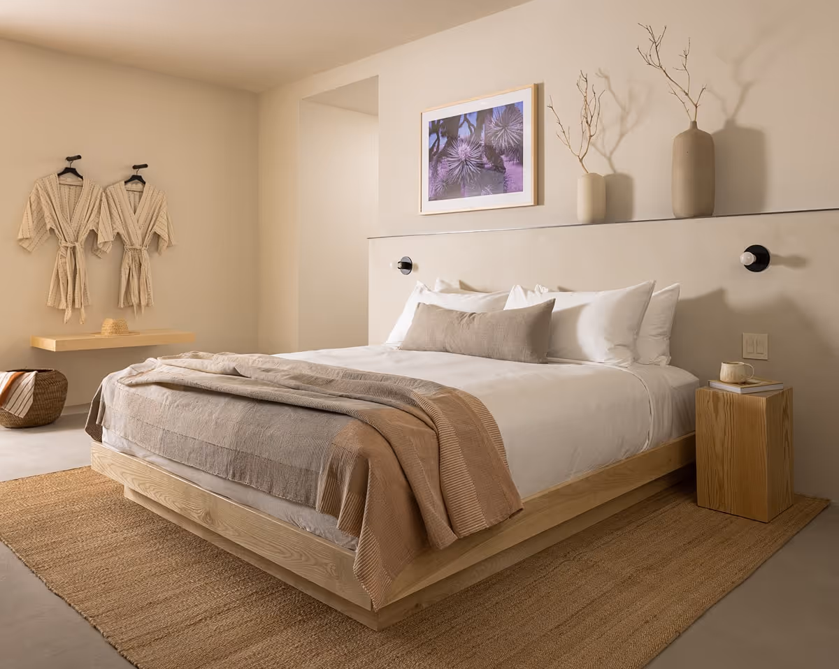 Neutral-toned bedroom featuring a low wooden platform bed with layered beige linens, wall-mounted robes, woven basket, and earthy décor accents.