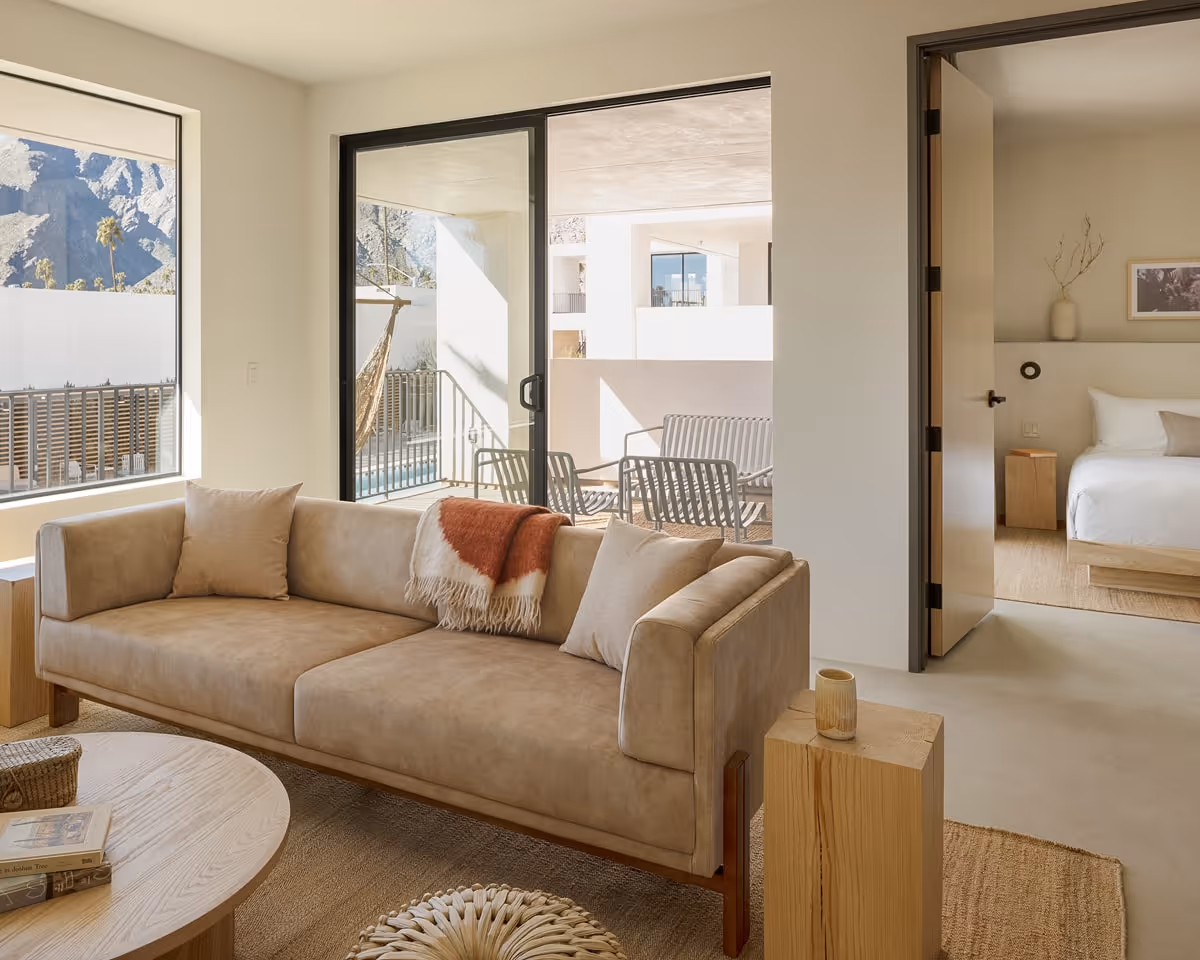 Modern living room with neutral sofa, wooden coffee table, mountain views through large windows, and access to bedroom and balcony.