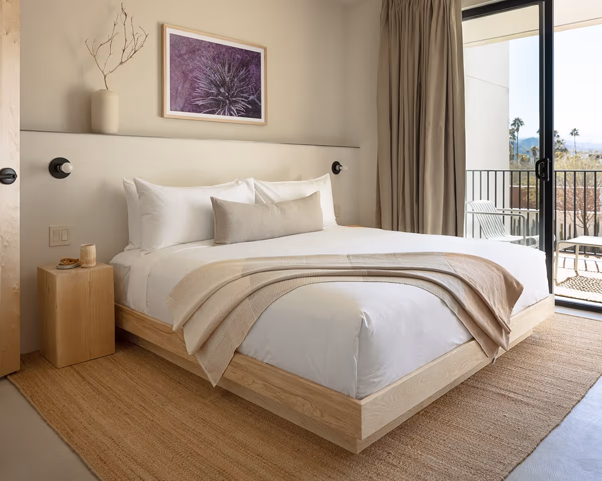 Minimalist bedroom with natural wood platform bed, soft neutral bedding, balcony access, and desert-inspired artwork.