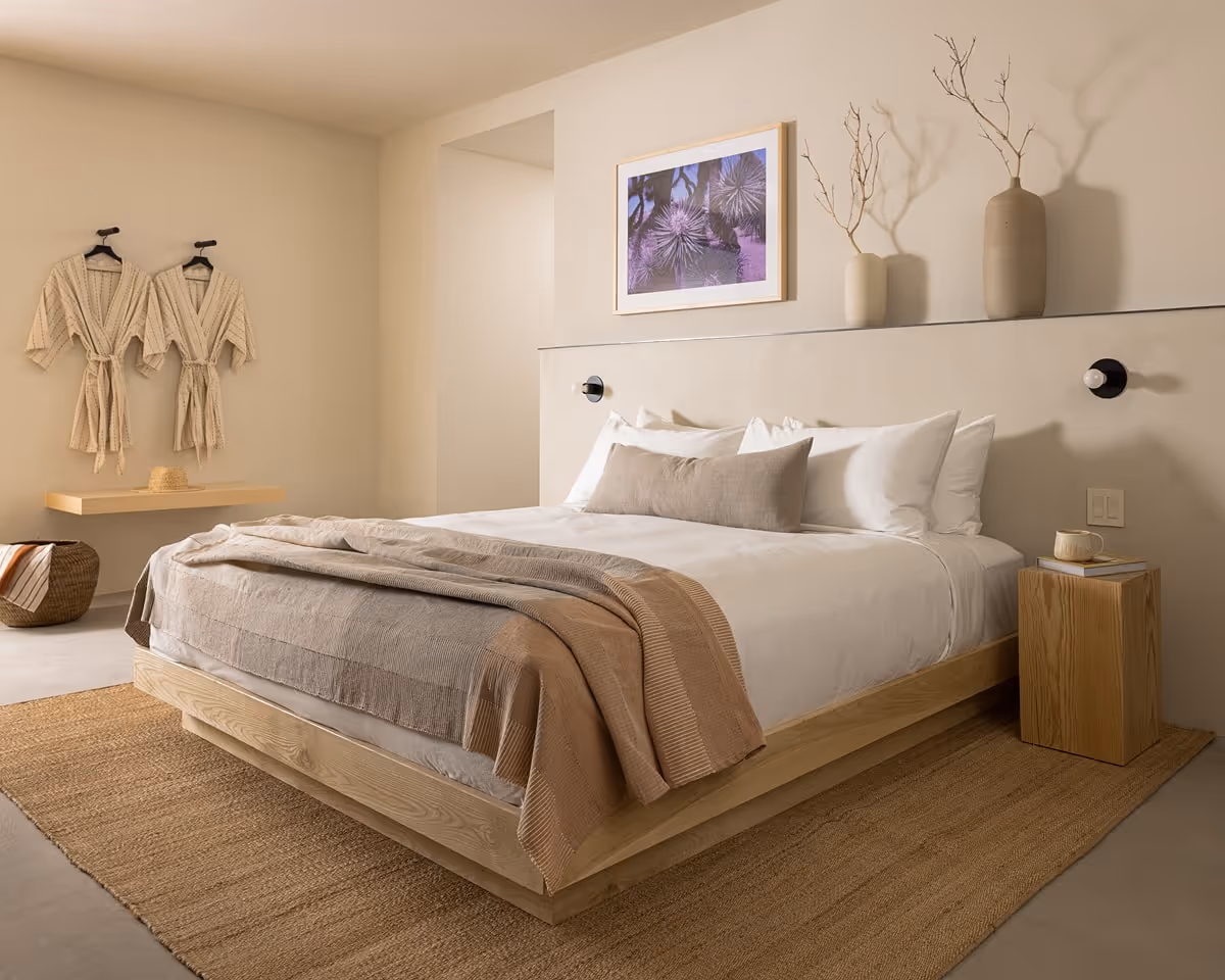 Minimalist bedroom with natural wood platform bed, neutral linens, woven rug, and wall décor in a modern suite.