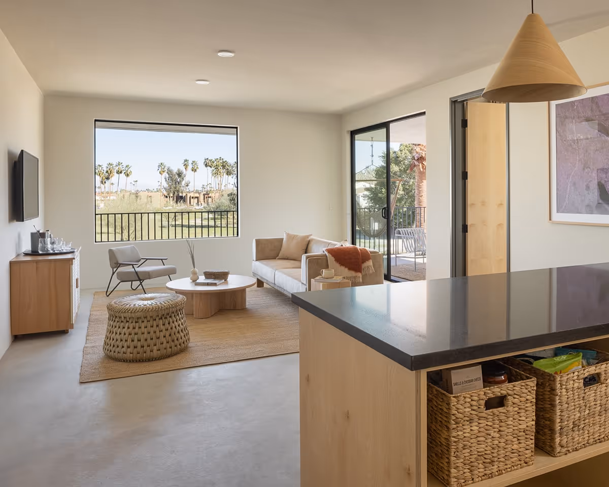 Spacious living room with modern neutral décor, large windows, natural wood furniture, and balcony views of palm trees.