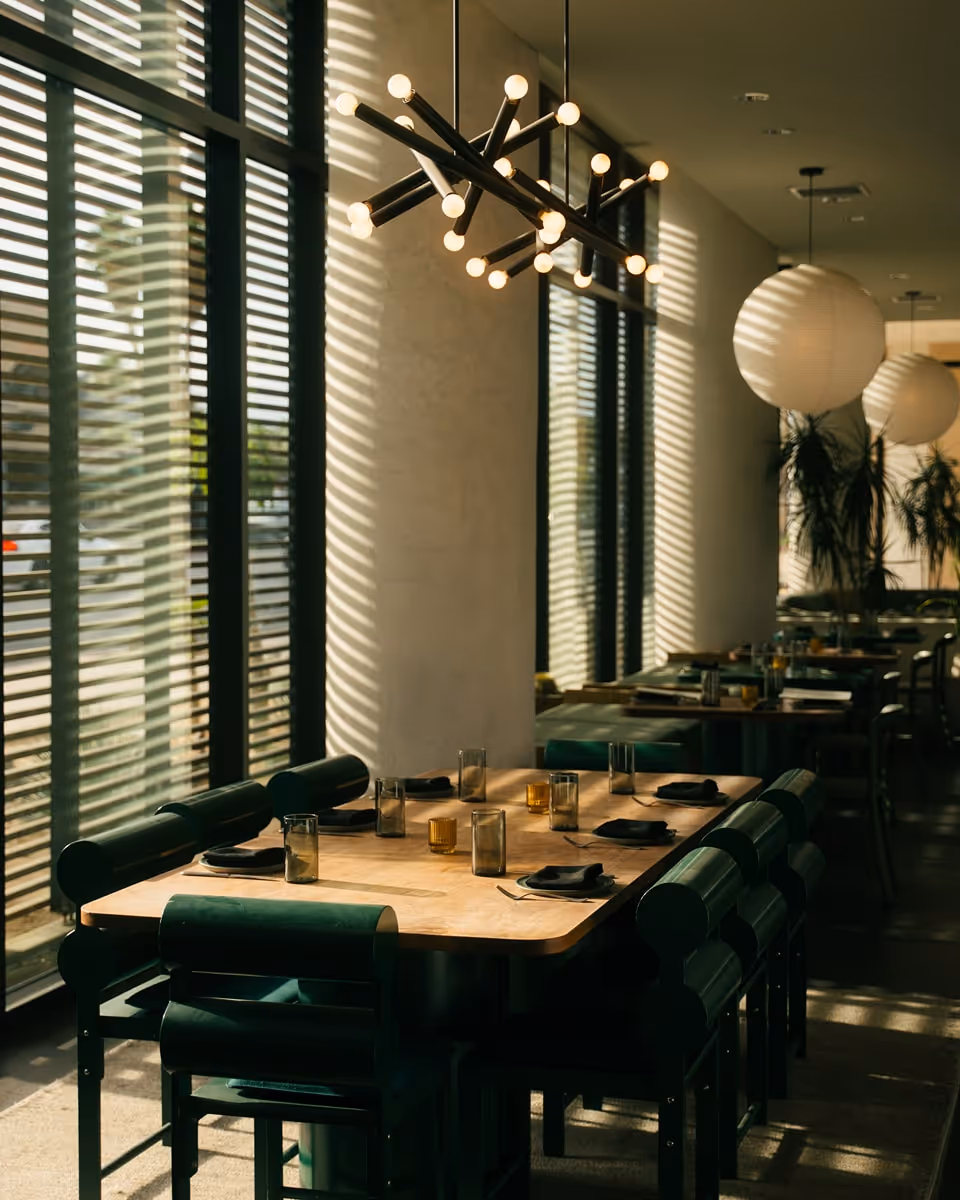 Modern restaurant dining area with wooden tables, dark green chairs, and statement lighting fixtures by large windows.