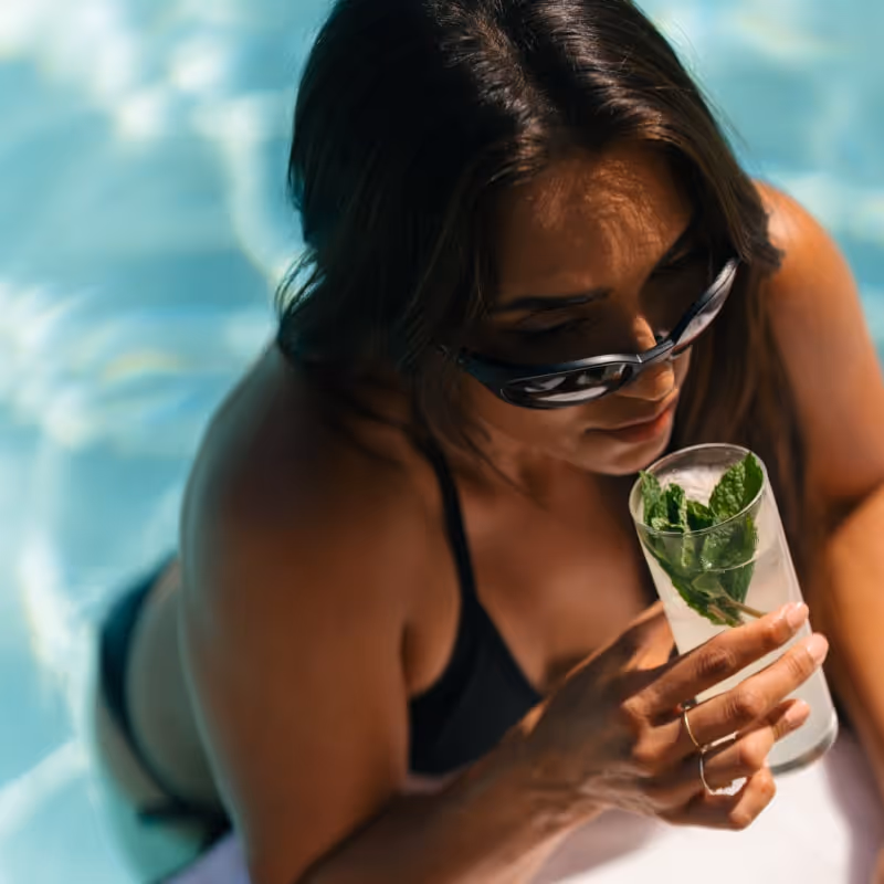 Woman relaxing poolside in sunglasses, enjoying a refreshing cocktail with mint garnish.