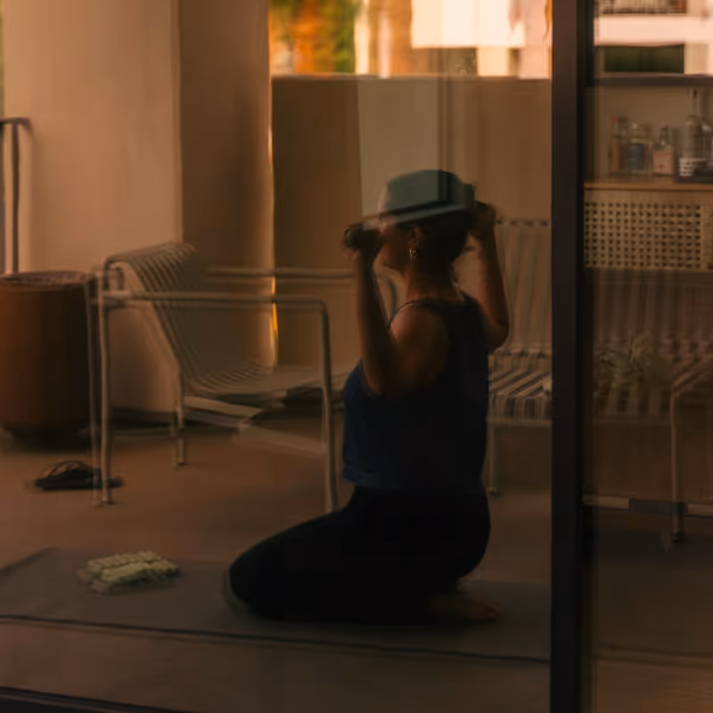 Woman exercising indoors with dumbbells on a yoga mat, reflected through a glass door.