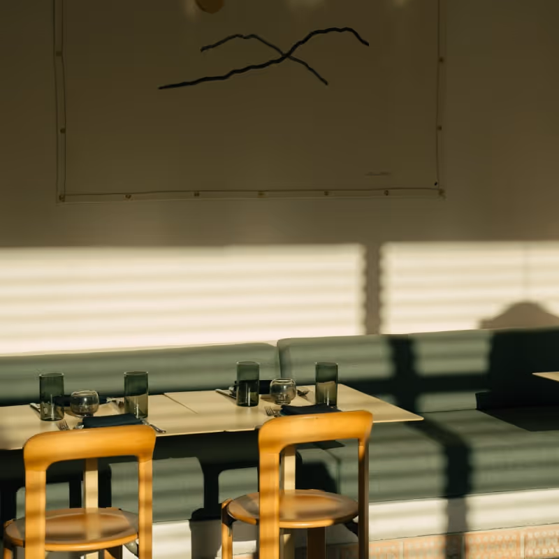 Minimalist restaurant interior with sunlight casting shadows on green bench seating, wooden chairs, and neatly set dining tables with glassware.