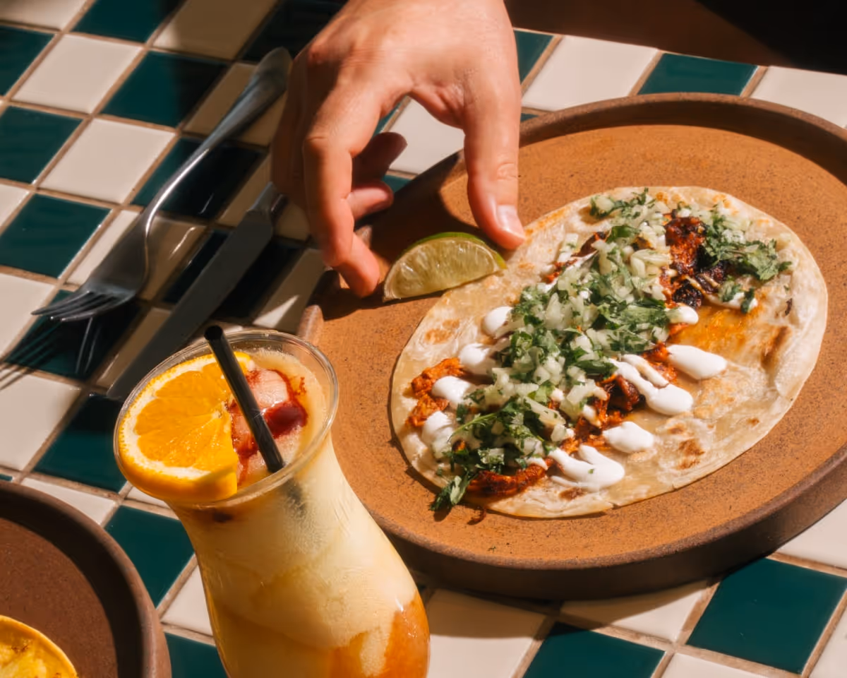 Hand reaching for a taco with cilantro, onions, and crema served with lime, paired with a citrus cocktail garnished with orange slice.