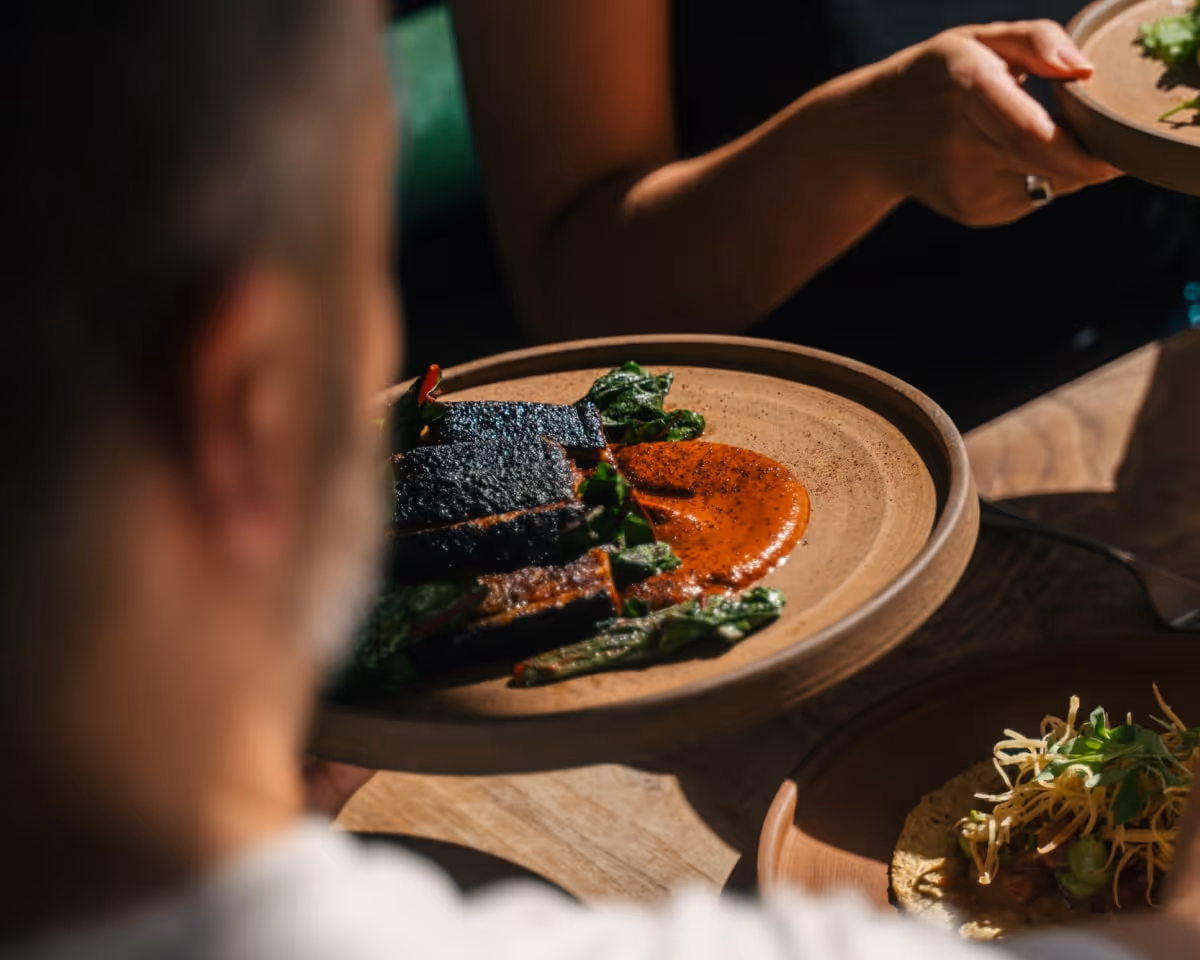 Close-up of a charred meat dish with roasted greens and rich red sauce served on a rustic ceramic plate in a restaurant setting.