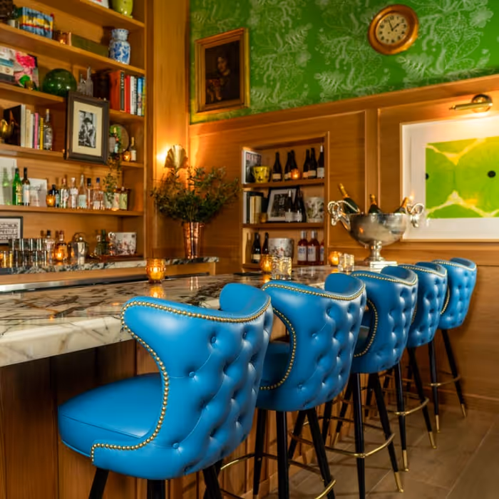Bar Cecil interior with blue tufted bar stools, marble bar, wood shelving and green wallpaper.