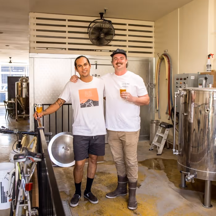 Las Palmas Brewery brewhouse with two brewers holding pints beside stainless steel tanks.