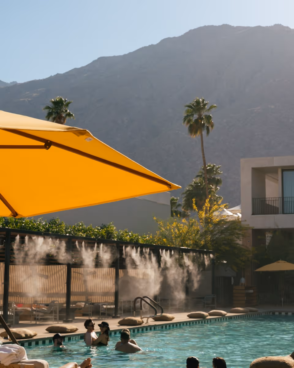 Hotel pool with guests swimming under yellow umbrellas and mist cooling system in Palm Springs