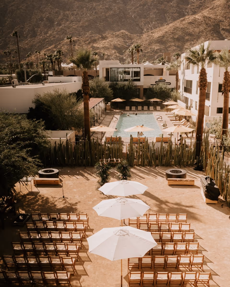 Desert resort wedding ceremony setup with wooden chairs, white umbrellas, cactus garden, pool area, and mountain backdrop in