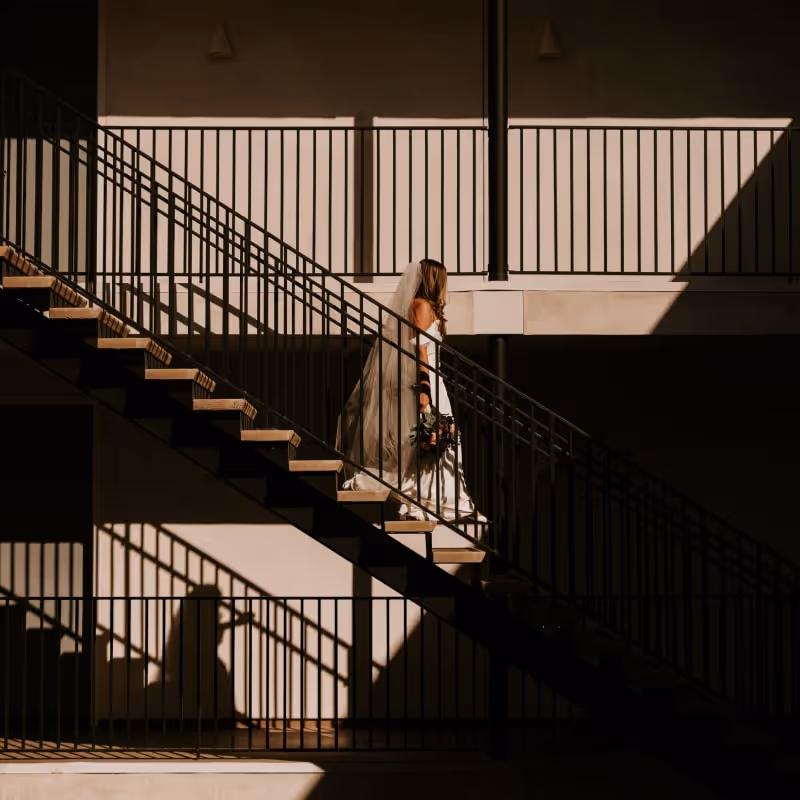 Bride in flowing white wedding dress descending modern outdoor staircase with dramatic shadows and geometric metal railings