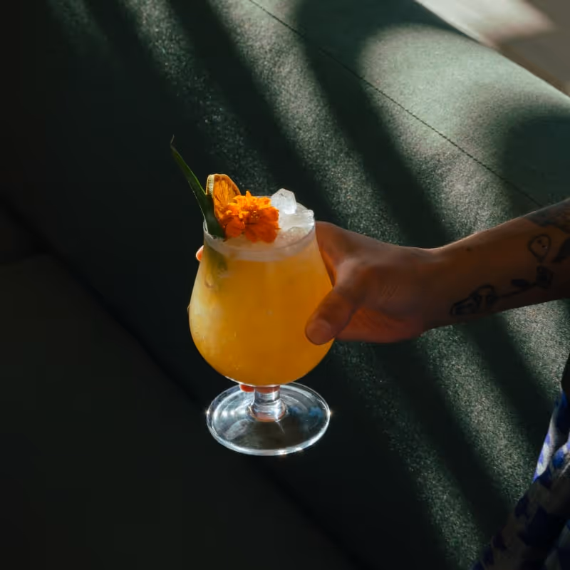 Hand holding a tropical cocktail with ice and orange garnish in natural sunlight