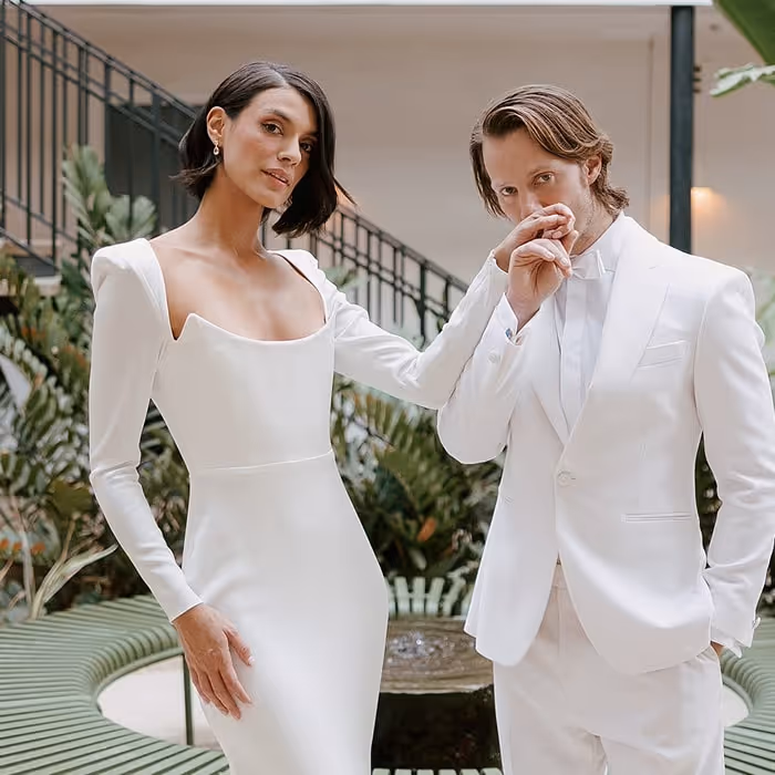 Stylish bride in modern long-sleeve wedding gown and groom in white tuxedo posing elegantly indoors