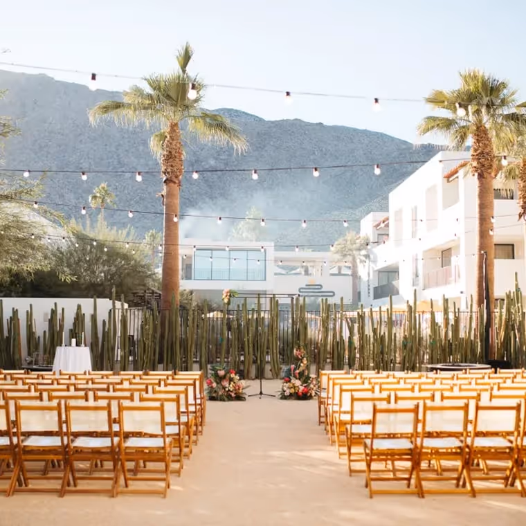 Outdoor wedding ceremony setup with bamboo chairs, cactus backdrop, palm trees, and string lights against mountain view
