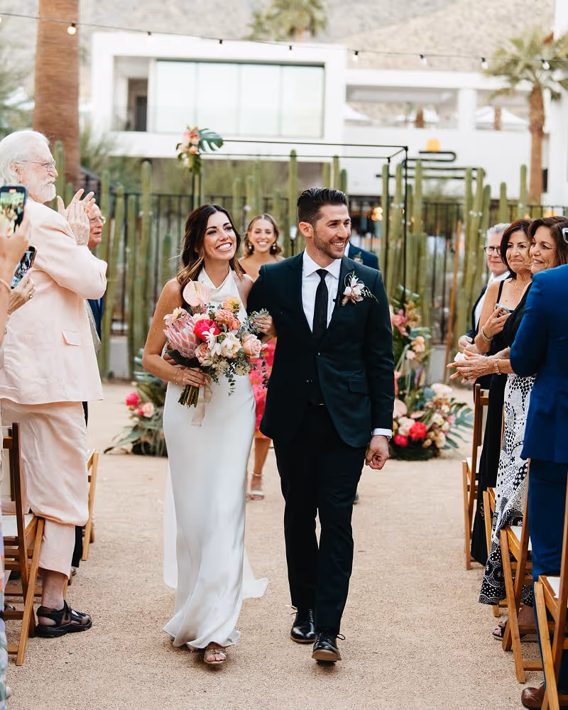Bride and groom walking down the aisle after ceremony, smiling and holding colorful bouquets while guests applaud