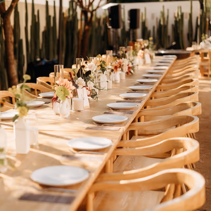 Long wedding reception table decorated with floral centerpieces, candles, and set white plates with wooden chairs