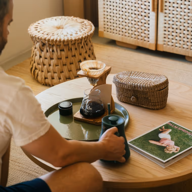 Guest enjoying pour-over coffee on a round wooden table with woven ottoman and Palm Springs magazine.