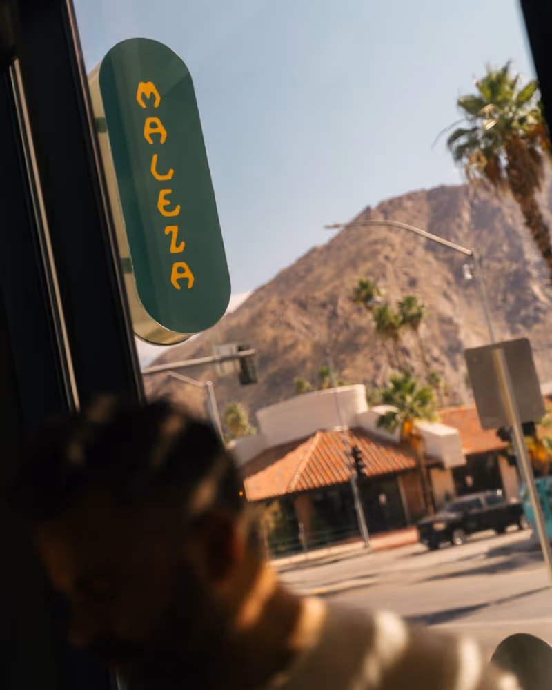 Maleza restaurant sign viewed through glass window with palm trees and mountains in the background.