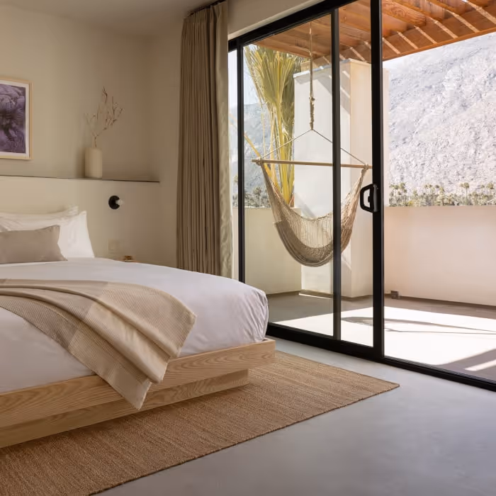 Minimalist hotel bedroom with natural wood bed, neutral decor, and balcony hammock overlooking desert mountains.