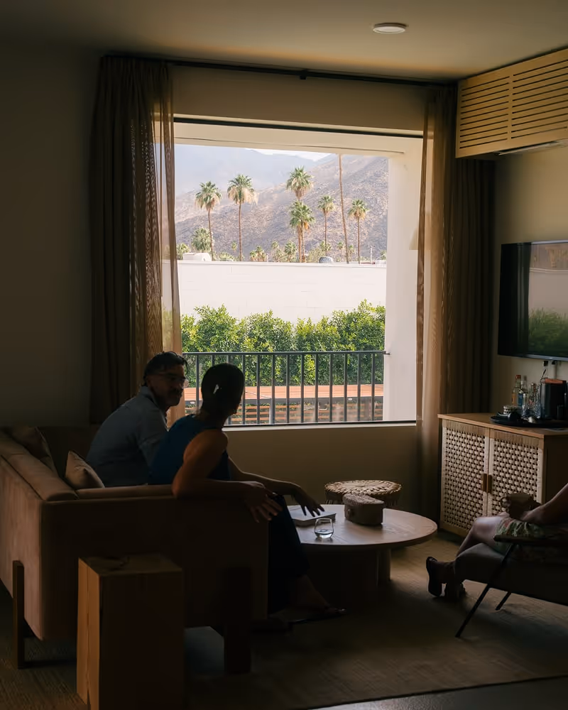 Couple sitting in hotel suite living area with large window view of palm trees and mountains.