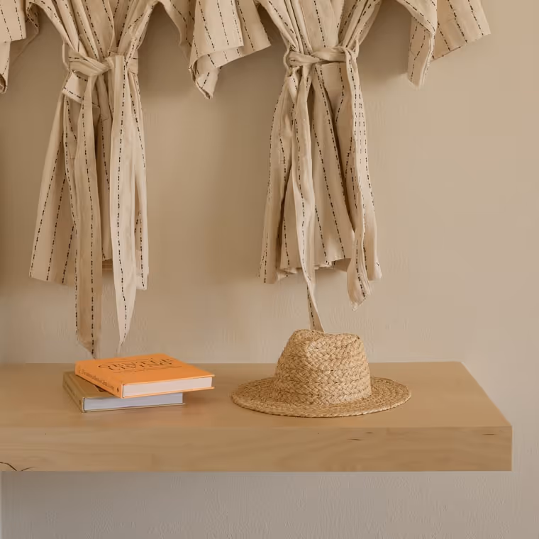Neutral-toned hotel robes hanging on wall with straw hat and books on wooden shelf.