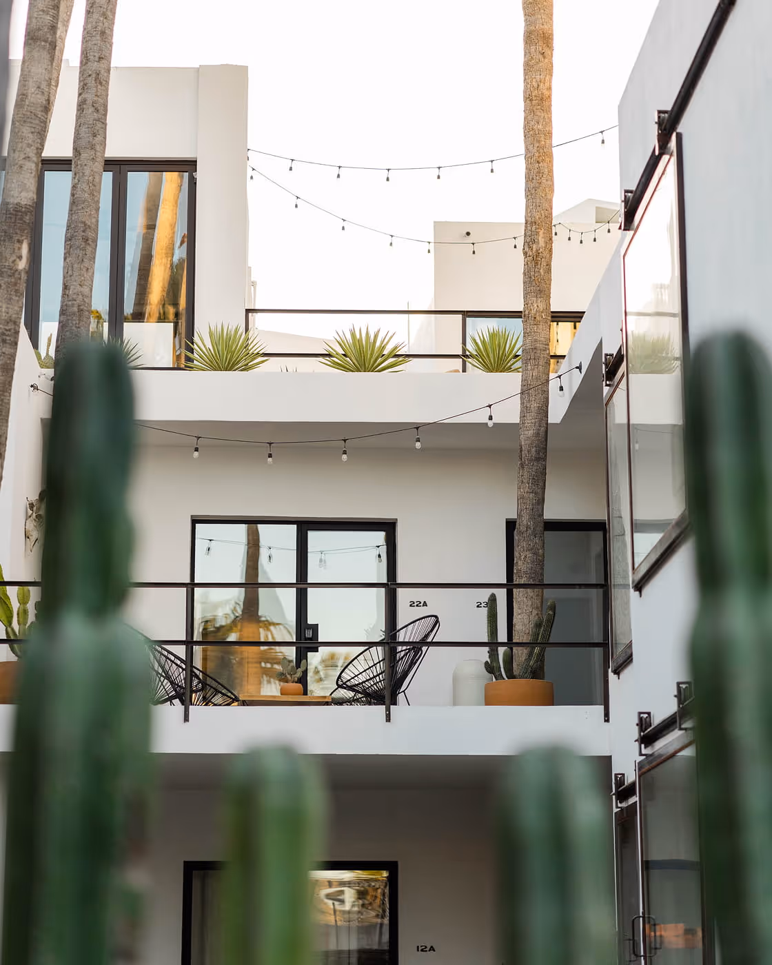Contemporary courtyard hotel with white stucco walls, black metal balconies, Acapulco chairs, cacti, and string lights in