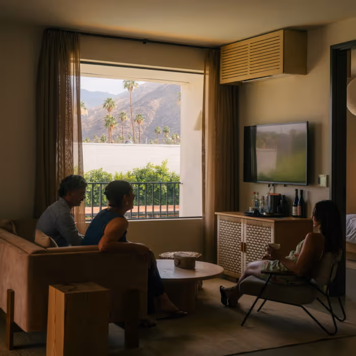 Friends relaxing in a modern hotel suite with a balcony view of palm trees and mountains.