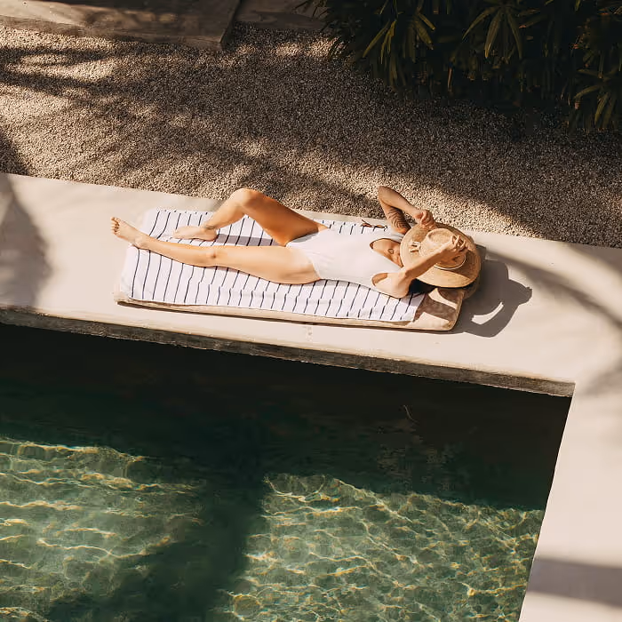 Woman in white swimsuit relaxing poolside on striped towel with straw hat, luxury tropical resort vacation setting