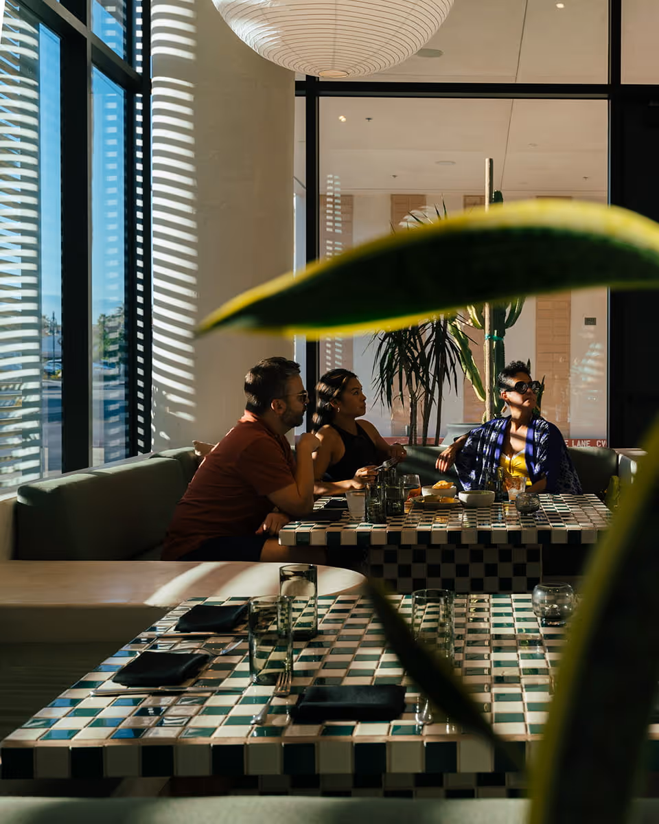 Three friends enjoying lunch at modern restaurant with checkered tile tables, tropical plants, and natural light streaming