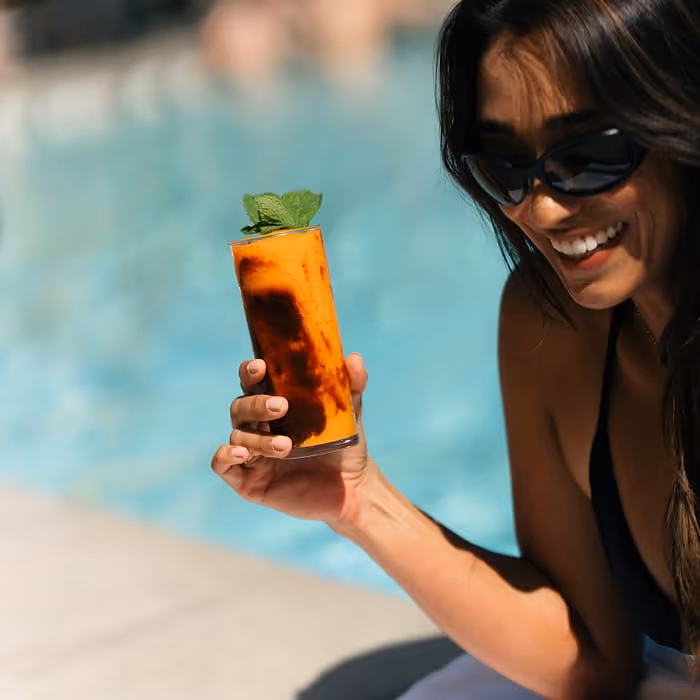 Smiling woman in black swimsuit holding orange mango cocktail with mint garnish by turquoise pool on sunny day