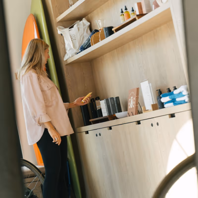 Woman browsing skincare products on wooden retail shelves next to colorful surfboards in coastal lifestyle shop