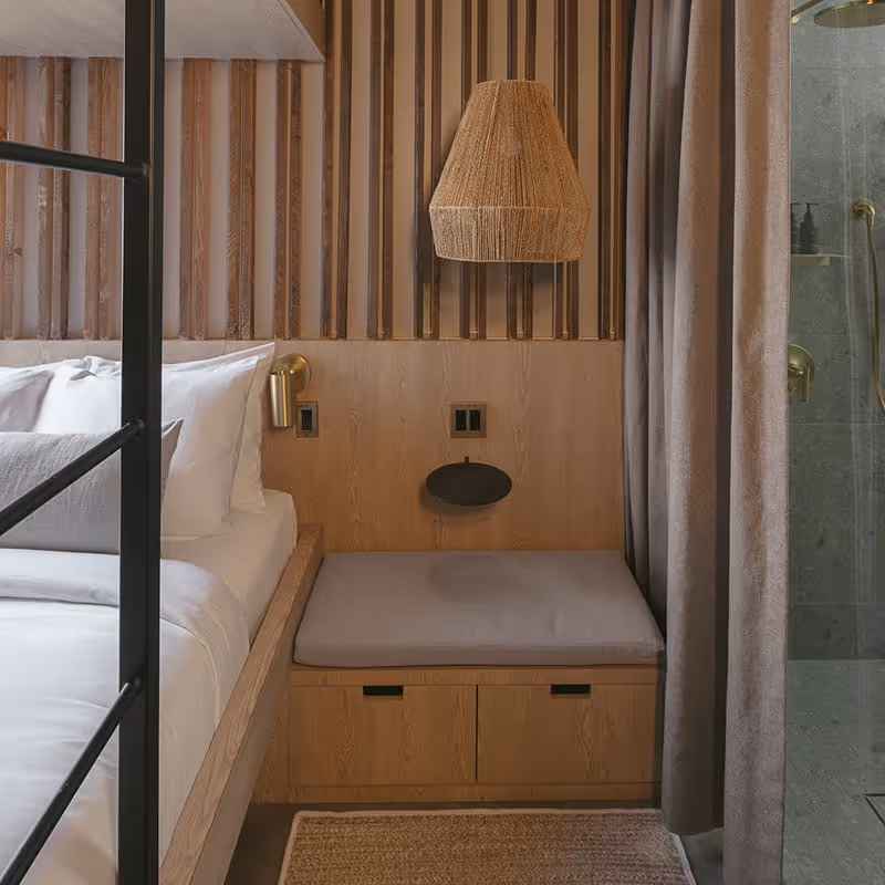 Modern boutique hotel bedroom with wood slat wall, built-in bench seating, woven pendant light, and glass shower with brass