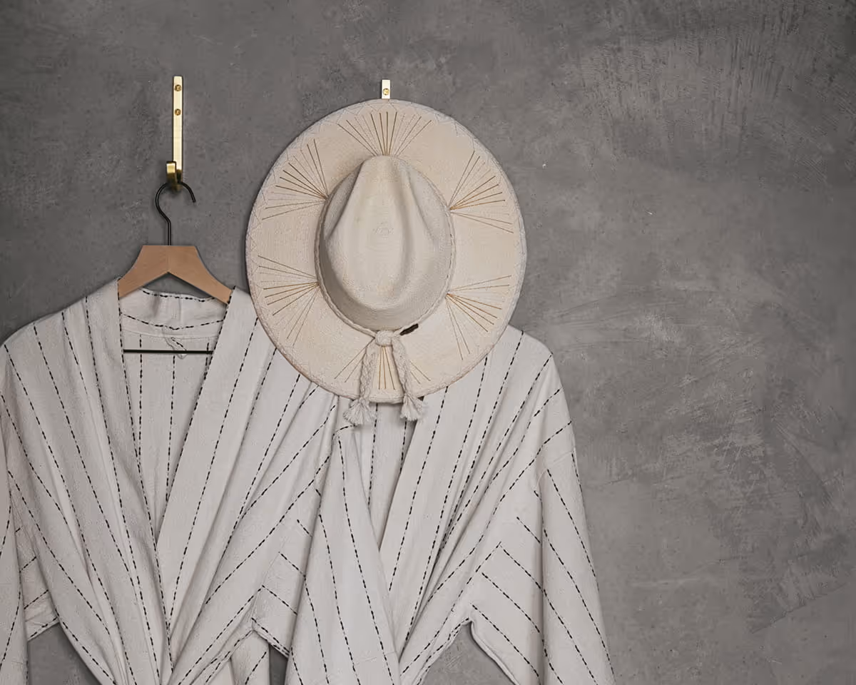 Cream striped linen robe on wooden hanger with straw sun hat against textured grey concrete wall in boutique hotel room