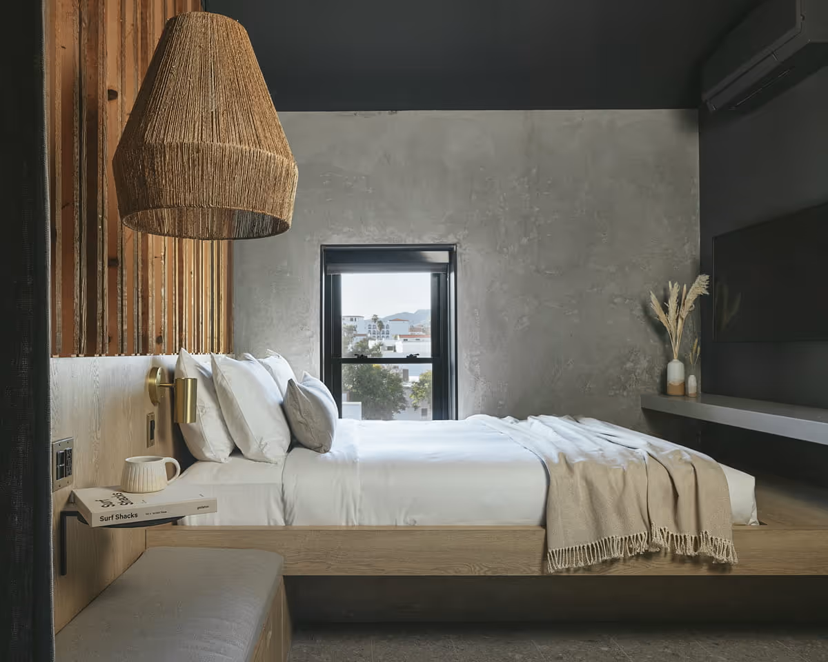 Contemporary boutique hotel room featuring platform bed, concrete walls, woven rattan pendant lamp, and Surf Shacks book on