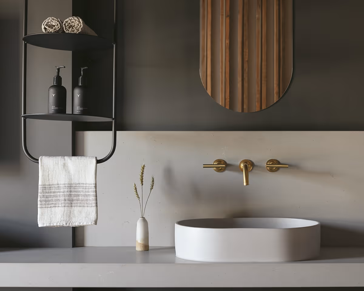 Minimalist bathroom vanity with oval vessel sink, brushed brass wall-mounted faucet, oval mirror, and wood slat accent wall