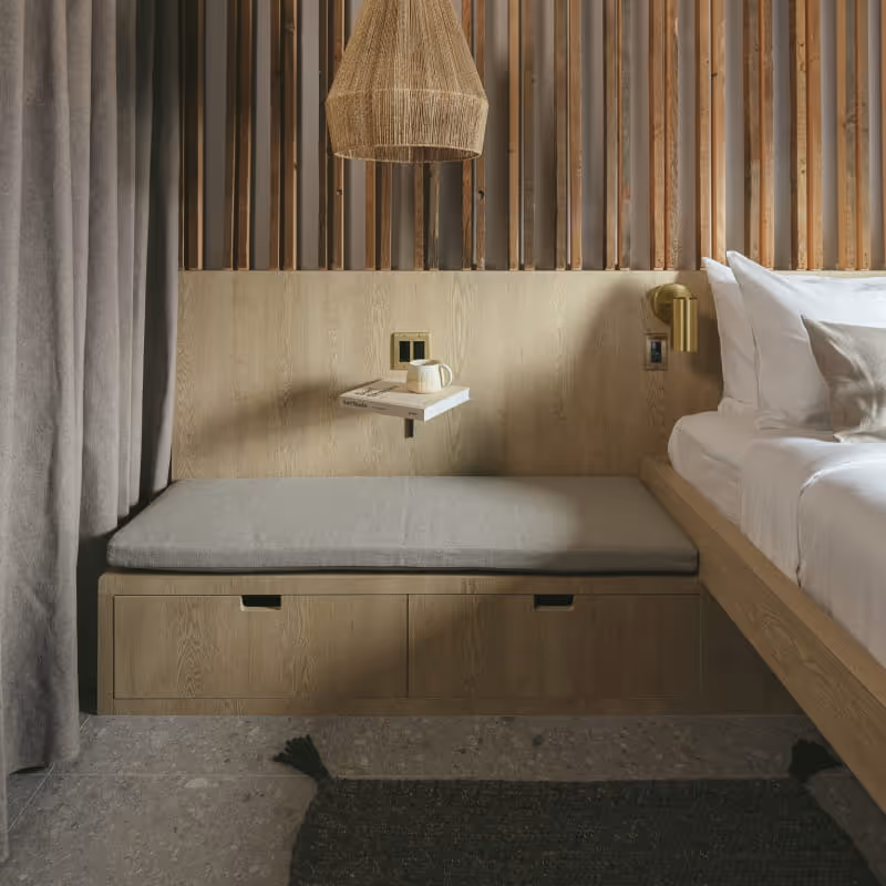 Modern hotel bedroom with oak wood platform bed, built-in storage bench, vertical wood slat wall, woven pendant light, and