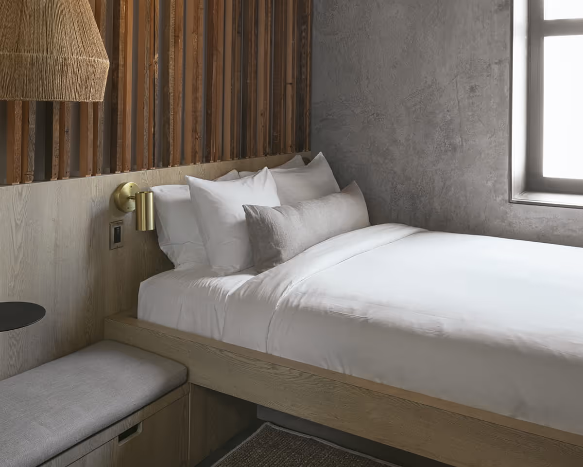 Minimalist boutique hotel room featuring white bedding on oak platform bed, textured concrete wall, wood slat headboard, and
