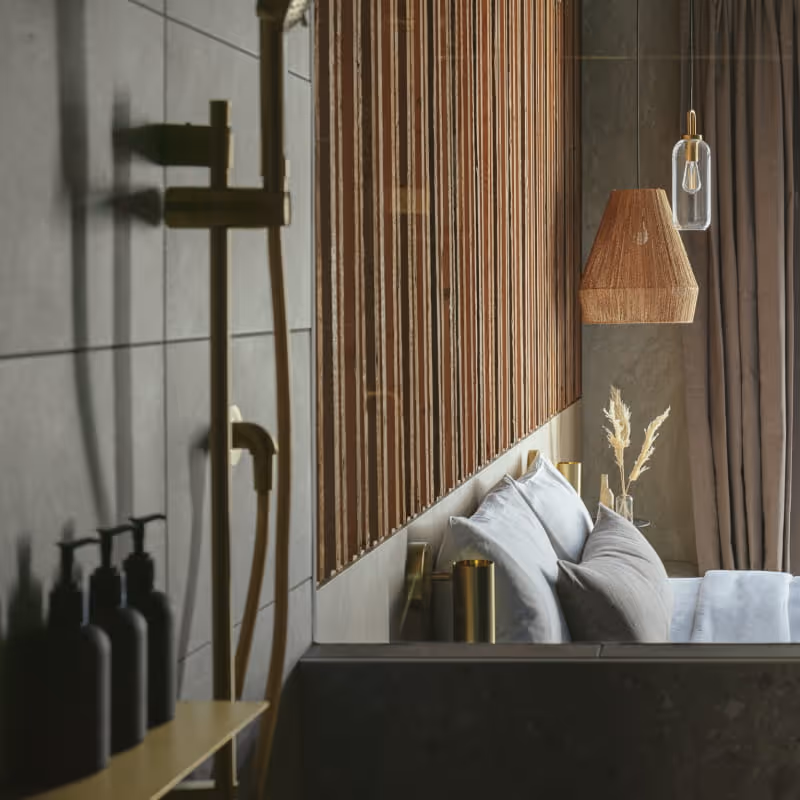 Modern hotel bathroom with wood slat accent wall, brass shower fixtures, and view into bedroom with woven pendant lamp