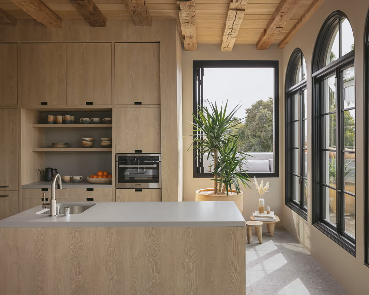 Modern rustic kitchen featuring light oak cabinetry, exposed beam ceiling, arched black-framed windows, and potted yucca