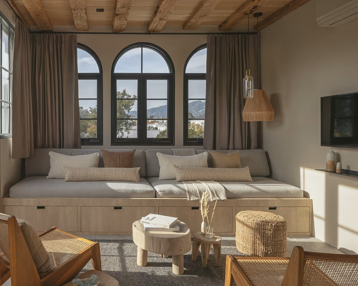 Cozy living room with exposed wooden ceiling beams, arched windows, built-in daybed sofa with storage, and woven rattan