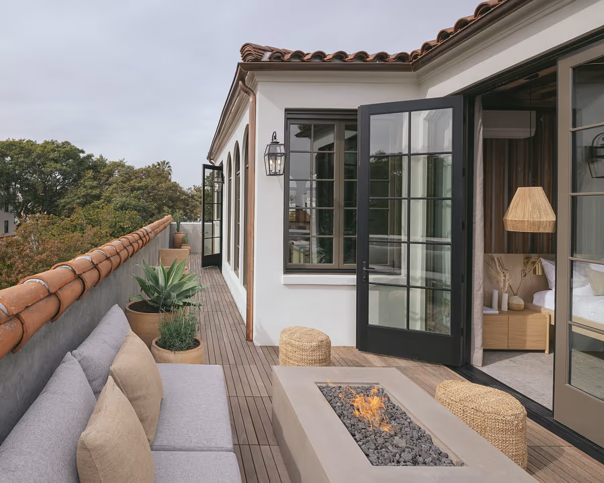 Spanish-style rooftop terrace with built-in gray sectional sofa, modern fire pit, woven poufs, and French doors leading to