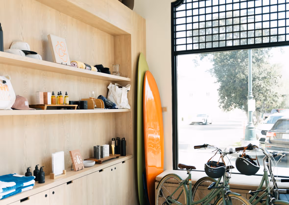 Coastal lifestyle boutique interior with wooden shelves displaying skincare products, surfboards, and vintage bicycles by